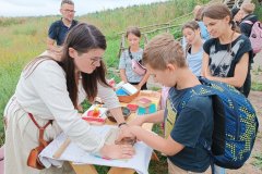 Na pierwszym planie dziecko odciskające na papierze drewnianą pieczątkę oraz pomagająca mu pracownica grodziska. Na drugim planie dzieci przyglądające się pieczątkom.