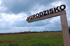 Na pierwszym planie drewniany kierunkowskaz z napisem „GRODZISKO” w tle widoczne zabudowania grodziska pośród łąk.