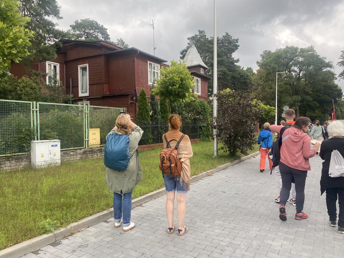 Grupa ludzi spacerujących po Kolumnie. W tle duży, drewniany dom oraz zielone drzewa.