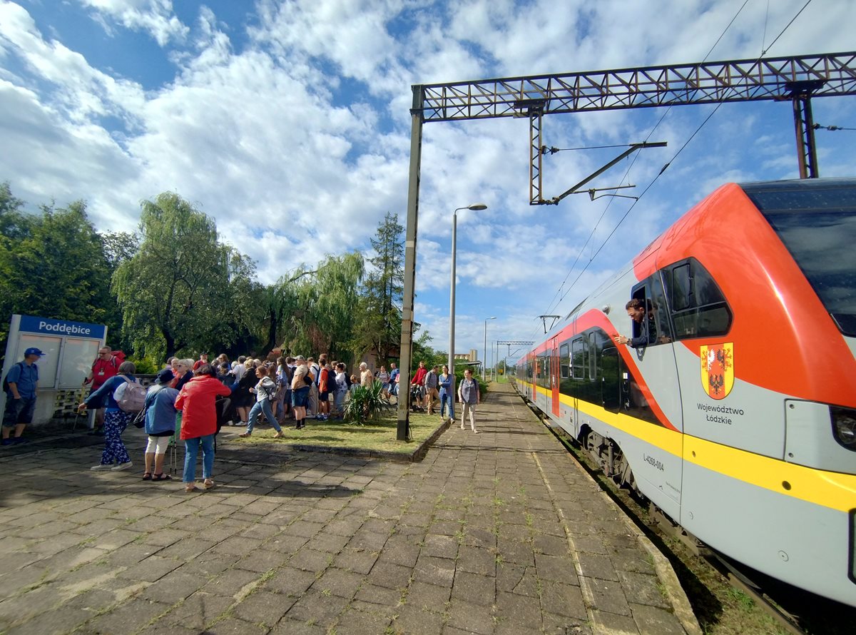 Grupa ludzi na stacji kolejowej w Poddębicach. Po prawej stronie czerwono-żółto-szary pociąg.