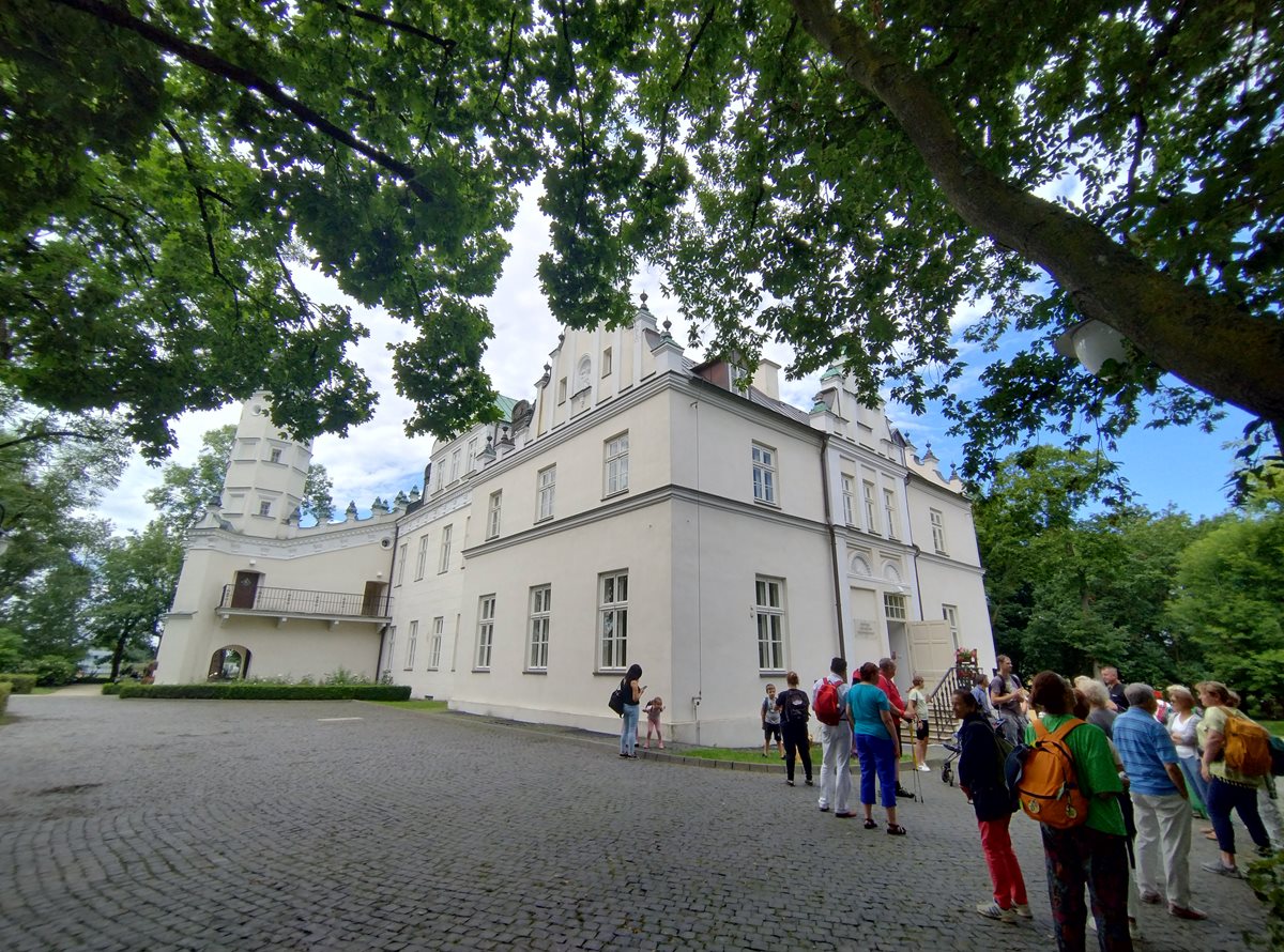 Po prawej strony znajduje się grupa ludzi stojąca przed Pałacem Grudzińskich. W centralnej części jest widok na pałac.