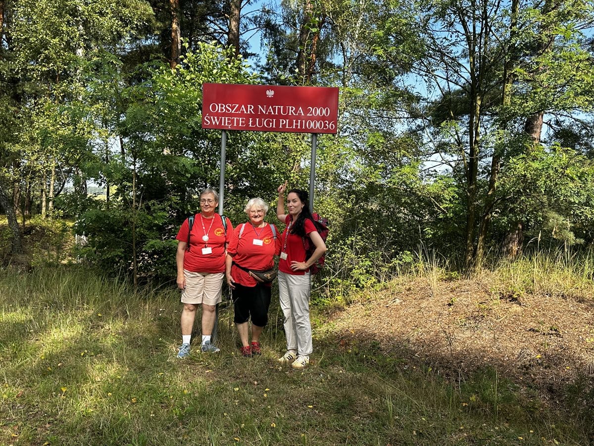 Trzy osoby (panie przewodniczki) stoją pod czerwoną tablicą z napisem Obszar Natura 2000 Święte Ługi PLH100036. W tle roślinność drzewiasta