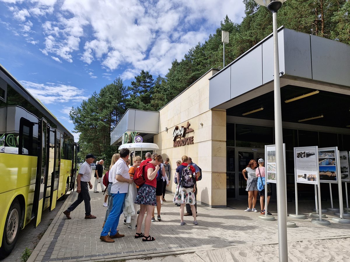 Grupa ludzi stoi przy budynku z napisem „Góry Nagórzyckie”. Po lewej stronie znajduje się żółty autobus.