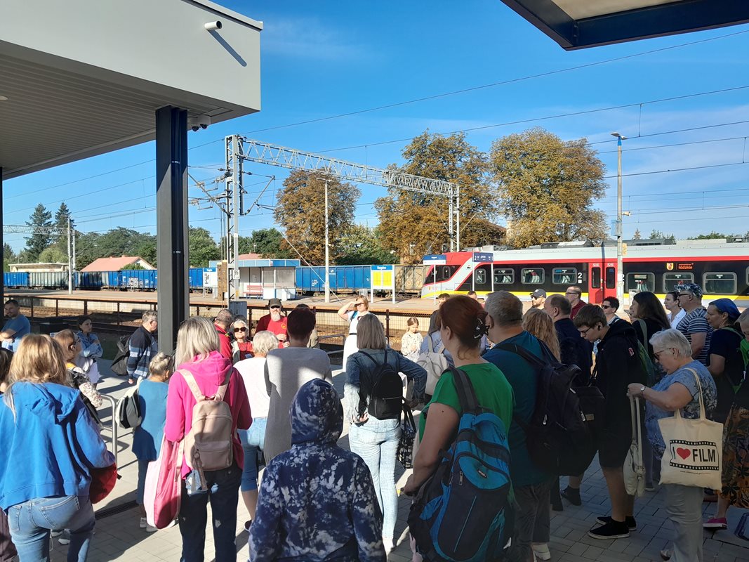 Grupa ludzi stojąca na chodniku, w tle widać tory kolejowe oraz czerwono-żółto-szary pociąg znajdujący się po prawej stronie obrazka