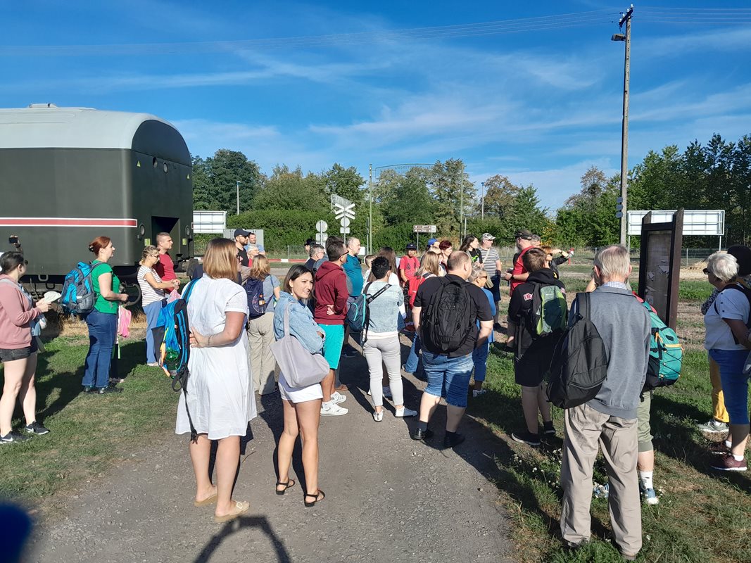 Grupa ludzie stojąca na chodniku oraz na trawie, za nimi widać wagon kolejki wąskotorowej, a w tle tory kolejowe