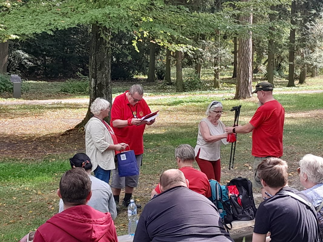 Na pierwszym planie przewodnicy w czerwonych koszulkach losują oraz wręczają kije do nordic walking uczestnikom wycieczki. W tle park oraz obserwujący losowanie wycieczkowicze. 